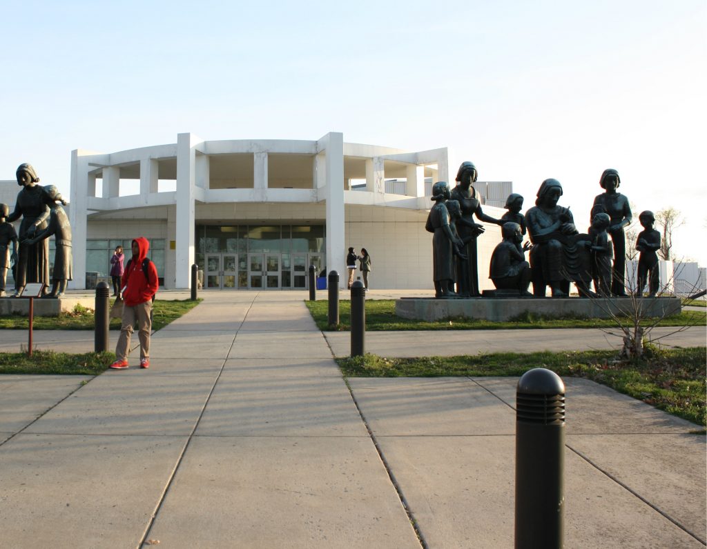 An off center view of the front of the school. On the right you can see a statue, on the left a child with a red hoodie. Up the path are the front doors of the school.